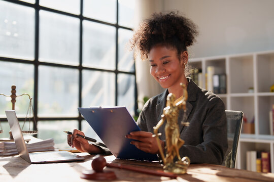 Young African American female lawyer sitting at her desk, reviewing and writing on a legal document with a pen, surrounded by law firm elements like the Gavel and Lady Justice