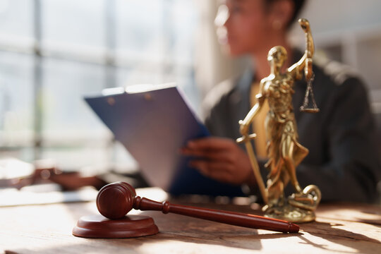 Lady Justice statue and a wooden gavel on a table, with a lawyer reviewing documents in the background, symbolizing legal proceedings and judicial system