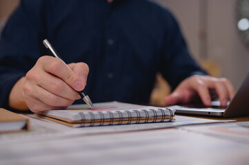 Businessman working in home office writing note to notebook while using laptop, taking notes from financial data and business reports in office environment. Productivity and planning concept.