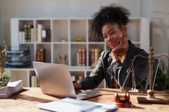 Young african american woman lawyer focused on her work, sitting at a wooden desk with a laptop, scales of justice, and gavel in a modern law firm office