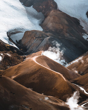View of a lone figure standing on a winding path atop a rugged, rusty-hued mountain range under a snowy glacier, steam rising from the earth, Kerlingarfj&Atilde;&para;ll, Highlands, Iceland.
