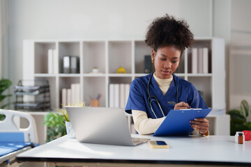 Female African American nurse sitting at a desk, reviewing patient information and writing notes on a clipboard with a laptop nearby in a modern clinic office