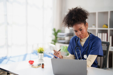 African American medical professional wearing scrubs and stethoscope, working on a laptop and...