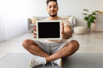 Young Arab guy showing tablet computer with empty screen while sitting cross legged on yoga mat at home, mockup for app or website design. Online sports advertisement concept