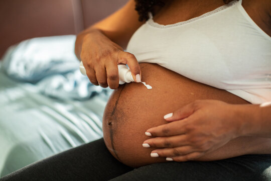 Calm adult woman applying lotion to pregnant belly at home