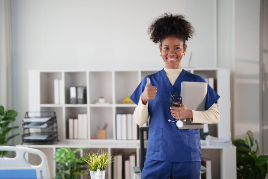 Female healthcare worker in blue scrubs giving a thumbs up gesture, smiling confidently and holding a laptop and stethoscope in a bright clinical or office environment