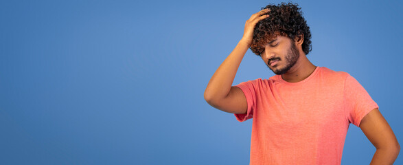 A man with curly hair stands against a blue backdrop. He has a serious expression on his face and is holding his head with one hand. The setting seems casual and indoor.