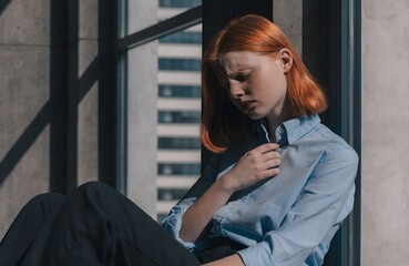 Urban Reflection Concept &ndash; Person in Light Blue Shirt with Hand on Collar Seated Near Window in Contemplative Pose, lifestyle, introspection, portrait, urban, window, shirt, red hair, concrete 