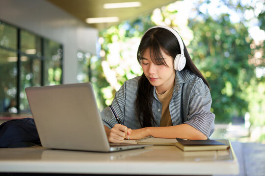 asian woman college student wearing headphone looking at laptop sitting at table in building