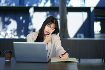Fototapeta premium Asian woman student freelancer talking on phone over book aside laptop sitting at table in building