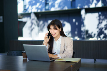 Asian woman student freelancer talking on phone over book aside laptop sitting at table in building