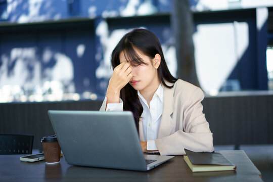 Focused young businesswoman looking at laptop pc computer screen having headache, migraine. Asian business woman holding hand near temples, feeling stressed and tired Sitting in the office building