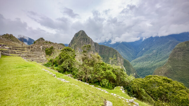 View of lush green terraces cascading down towards ancient stone ruins, crowned by a towering, cloud-kissed peak, shrouded in mist, Machu Picchu, Peru.