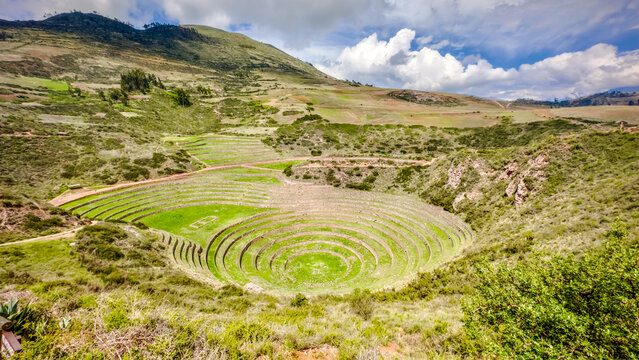 View of circular agricultural terraces cascading down into a verdant depression, framed by rugged hills under a bright sky, Moray, Peru.