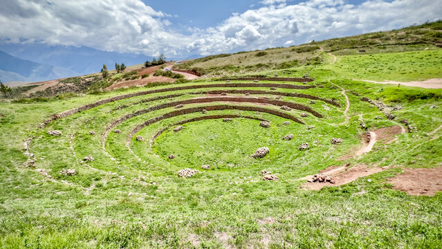 View of concentric agricultural terraces dropping into the earth, a vibrant green against the ancient stonework and cloudy sky, Moray, Peru.