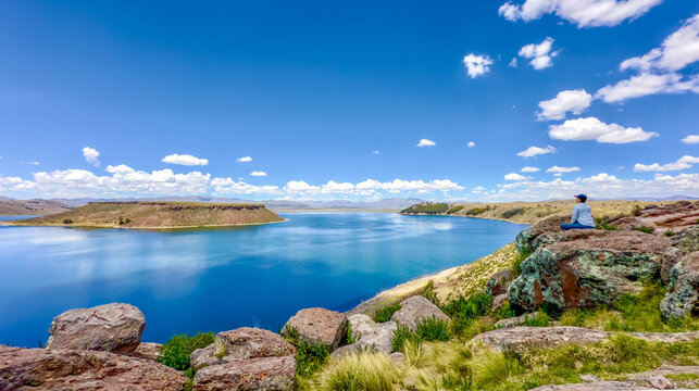 View of bright blue waters under a vast sky with fluffy clouds, contrasted by the rugged, rocky terrain and greenery, Sillustani, Peru.