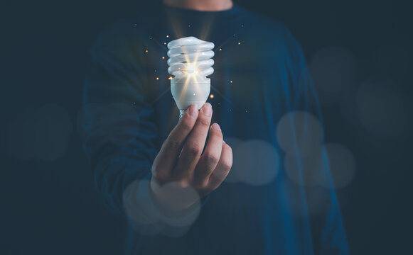 Hand holding glowing energy saving spiral light bulb on dark background, representing innovation, clean energy, sustainability, green technology, eco friendly ideas and future electricity concept.