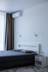 Light bedroom with bed near window and air conditioner above the bed. Bedroom interior.