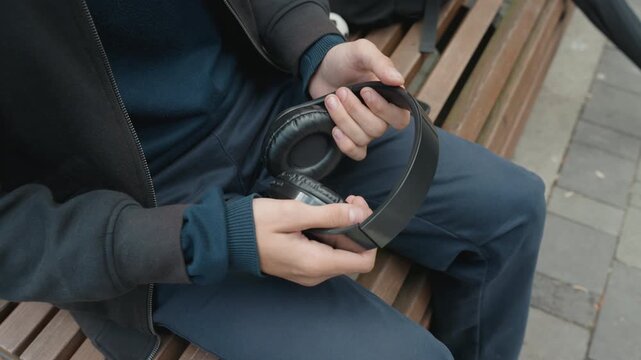 Man sitting on bench adjusting headphones, closeup of hands holding black overear headphones, casual jacket and sweater visible, city pavement background, subtle motion as he inspects earcup,
