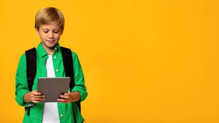 Obraz premium Smiling caucasian teen boy student with backpack typing on tablet and watching educational video, isolated on yellow background, copy space. Online education, remote schooling, distance learning class