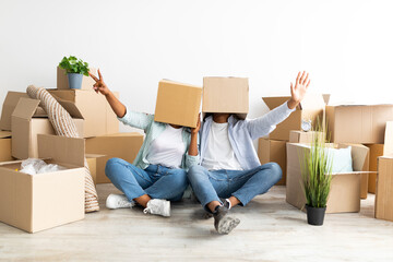 Joyful black couple with cardboard boxes on heads, sitting on floor and having fun, celebrating moving day, relocation to new apartment. Home, people, moving and real estate concept