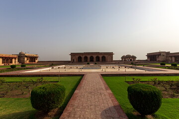 View of symmetrical gardens lead to aged buildings under a pale sky, with contrasting textures of brick and manicured lawns, Lahore, Punjab, Pakistan.