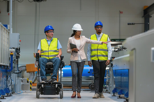 A female manager guides male factory workers and a worker with disability in a wheelchair through production operations in a manufacturing plant.
