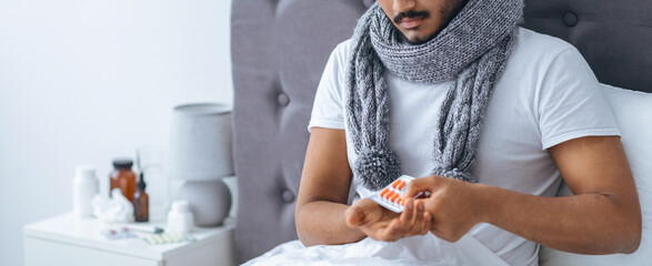 A man rests in bed while wearing a scarf. He holds a blister pack of medication, indicating he is...