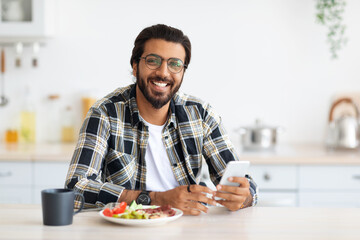 Cheerful middle-eastern young man with long hair and stylish beard in casual enjoying breakfast,...
