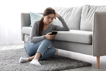 Upset frustrated pretty caucasian millennial lady suffering from depression sitting on floor and reading message at home. Human face expression, emotion, bad news reaction, device addiction and bored