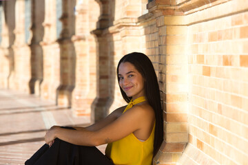 A young, pretty latin woman wearing a yellow shirt and black trousers is sitting on the floor with her back against the wall. She has her hands on her knees and looks happy and smiling.