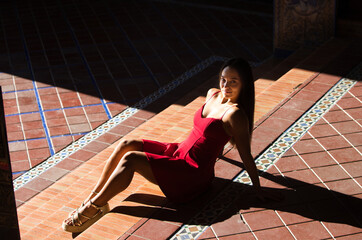Spanish woman, with long dark hair, young and pretty, dressed in red, sitting on the ground in a sunny spot and looking up. The photo is taken from above.