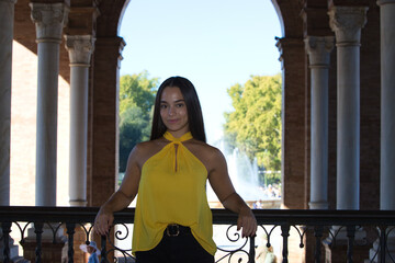 A young, dark-haired, pretty Latin woman wearing a yellow shirt and black trousers, leaning with her elbows on the railing of the Plaza de Espa&ntilde;a in Seville. The woman is happy.