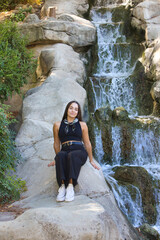 A young, pretty Spanish woman with dark hair sitting on the rocks next to the waterfall in the park. The woman is wearing black jeans and a black T-shirt with a scarf around her neck.