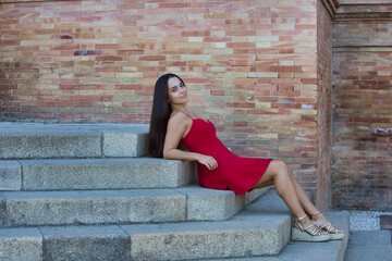 A young, pretty Spanish woman with long dark hair, dressed in red, is sitting relaxed on the steps of the staircase in Plaza Espa&ntilde;a in Seville, Spain.