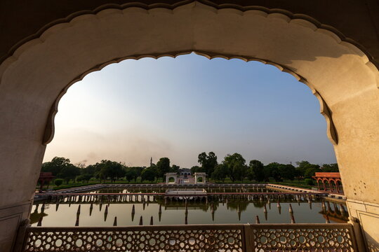 View of a serene pool reflecting trees and a distant pavilion framed by an ornate archway, creating a tranquil scene, Lahore, Punjab, Pakistan.