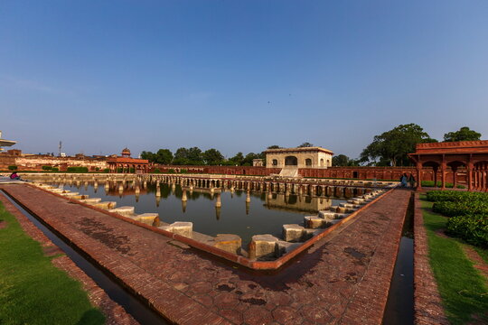 View of tranquil pool reflecting architecture under a clear sky, bordered by red brick pathways and lush greenery, Lahore, Punjab, Pakistan.