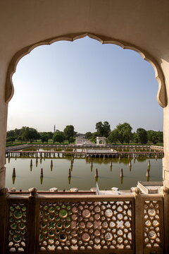 View of a serene pond reflecting the clear sky, framed by an ornate archway and decorative stone railing, creating a peaceful scene, Lahore, Punjab, Pakistan.