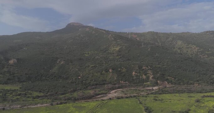 Aerial view of a large mountain covered with vegetation and a flat top, with green fields at its base under a cloudy sky, Pollina, Sicilia, Italy.
