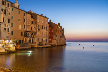 View of aged buildings glow with warm light against the serene Adriatic sea as twilight paints the sky in soft hues, Rovinj, Istria County, Croatia.