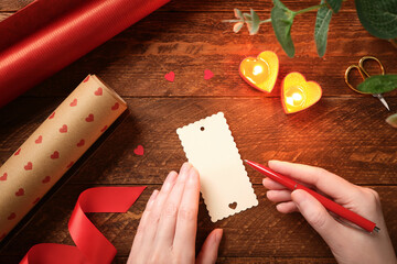 Romantic flat lay with hands writing gift tag on wooden table. Heart candles, red ribbon, wrapping paper. Handmade greeting with copy space