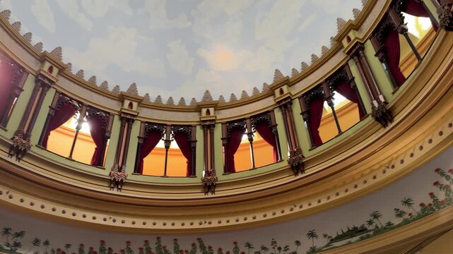 An ornate rotunda shows a circular dome with arches, columns, and rich curtains. Decorative molding and painted ceiling details highlight classic architecture.