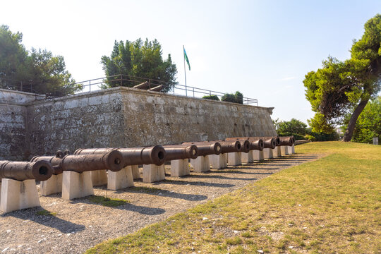 View of aged cannons lined up with precision against the backdrop of a stone fortification, the warm hues contrasting with the green flag, Pula, Istria County, Croatia.
