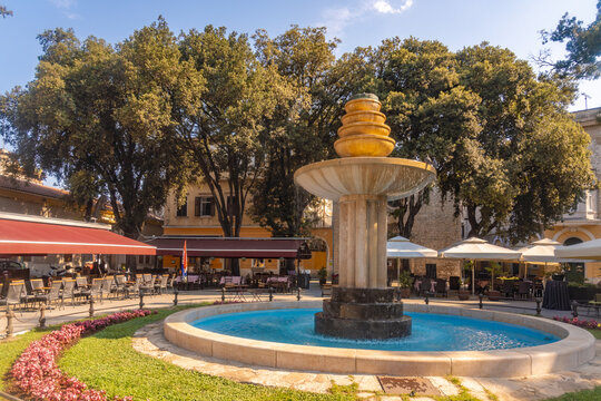 View of a vibrant turquoise fountain shimmering under the sun, embraced by lush greenery and shadowed cafes, in a picturesque European square, Pula, Istria County, Croatia.