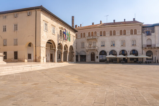 View of creamy stone buildings define the square, arches framing shadows, flags fluttering gently under the clear sky, in Pula, Istria County, Croatia.