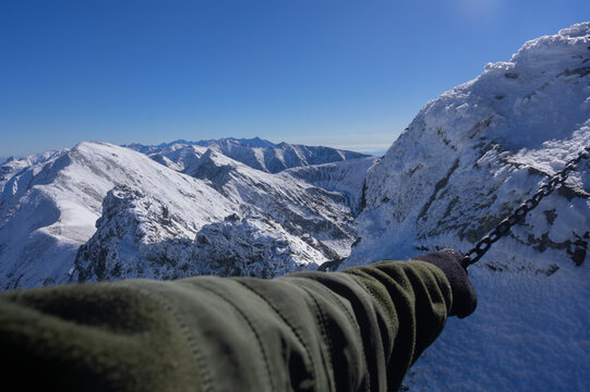 View of snow-capped peaks under a clear blue sky, a climber's gloved hand gripping chains on the mountainside, Banikov, Zilina Region, Slovakia.