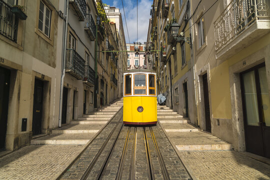 View of the bright yellow funicular ascends the steep cobbled street, framed by aged buildings under a blue sky with fluffy clouds, Lisbon, Lisbon, Portugal.