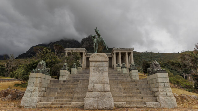 View of powerful bronze statues atop stone steps, framed by imposing architecture and a dramatically clouded mountain, Rhodes Avenue, Cape Town, Western Cape, South Africa.