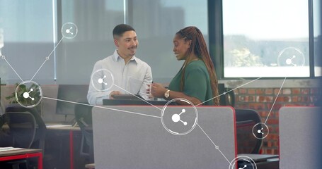 Talking colleagues in green blouse and light shirt at laptop by partition in office, network icons