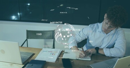Writing man in blue shirt leaning over notebook at wooden table, two laptops, globe, copy space